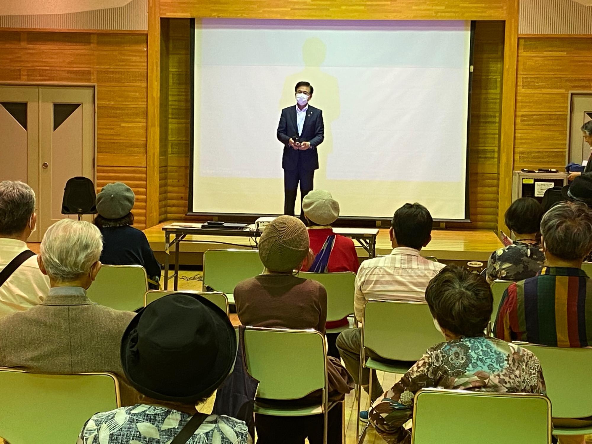 木目調の壁の部屋で、高橋市長がプロジェクタースクリーンの前に立って、椅子に座っている聴衆に向けて話している写真