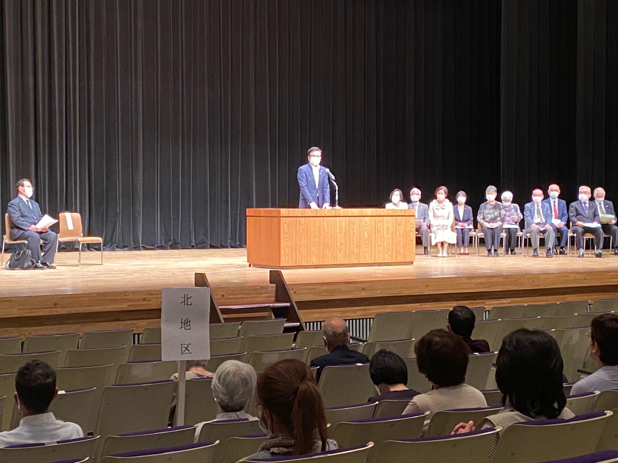 広いステージで、スーツを着てマスクを着用した高橋市長が演台に立ち、観客席に座る多くの人々に向かってスピーチをしている様子を捉えた写真