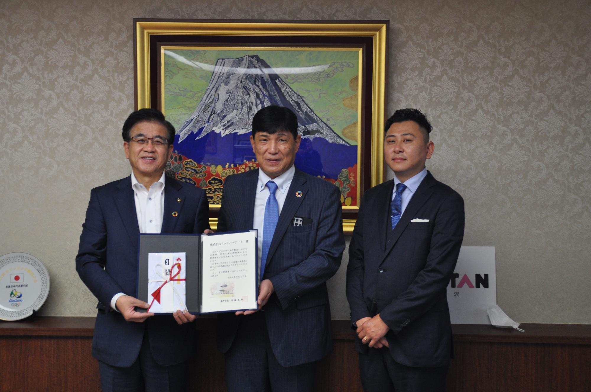 目録と証書ホルダーを一緒に持っている高橋市長と男性、その横に両手を前に組んでいる男性が並んで立ち、カメラに笑顔を向けて記念撮影に応じている写真