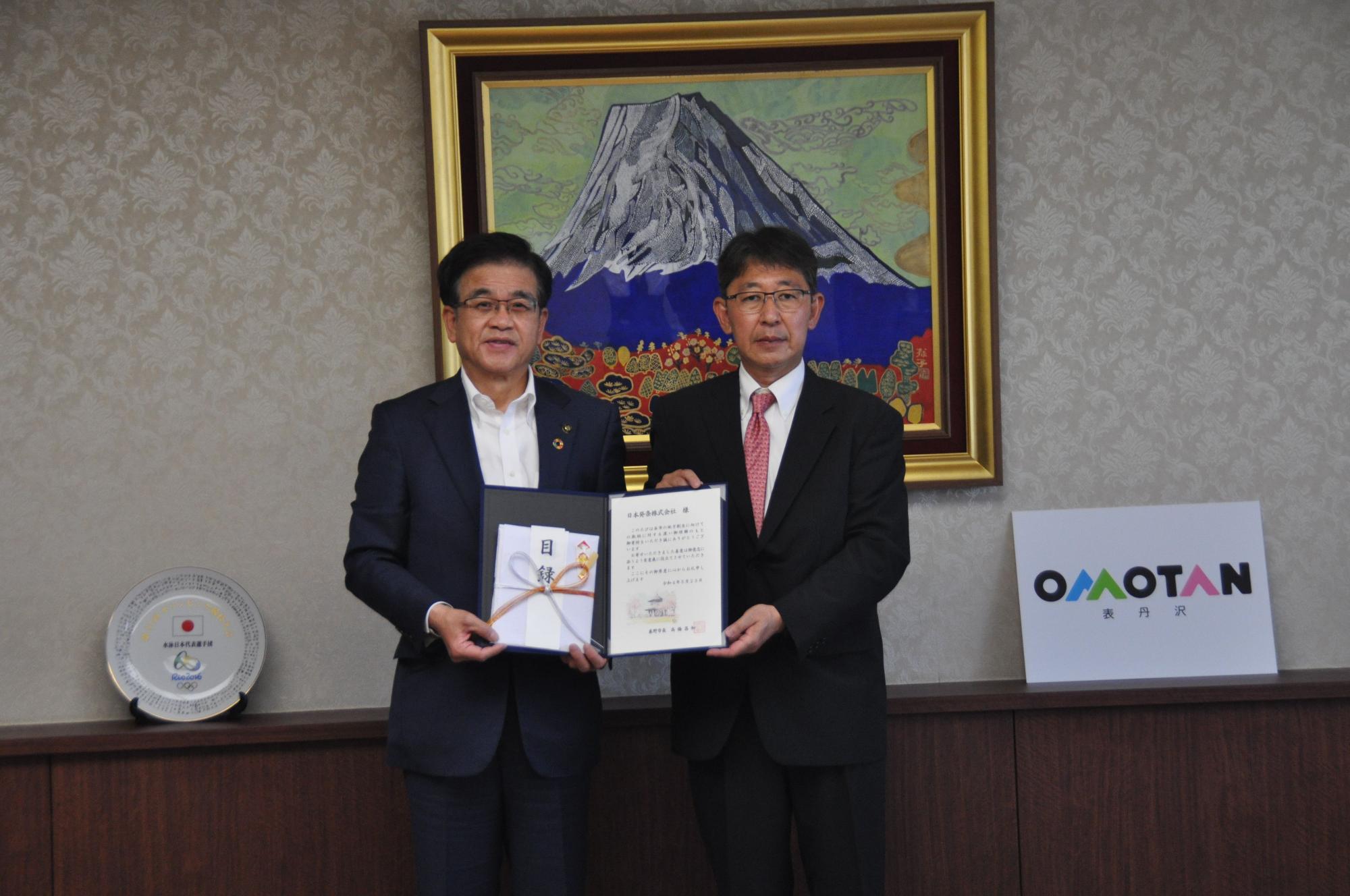 高橋市長と新藤取締役社長が一緒に目録と証書ホルダーを持ち記念撮影を行っている写真