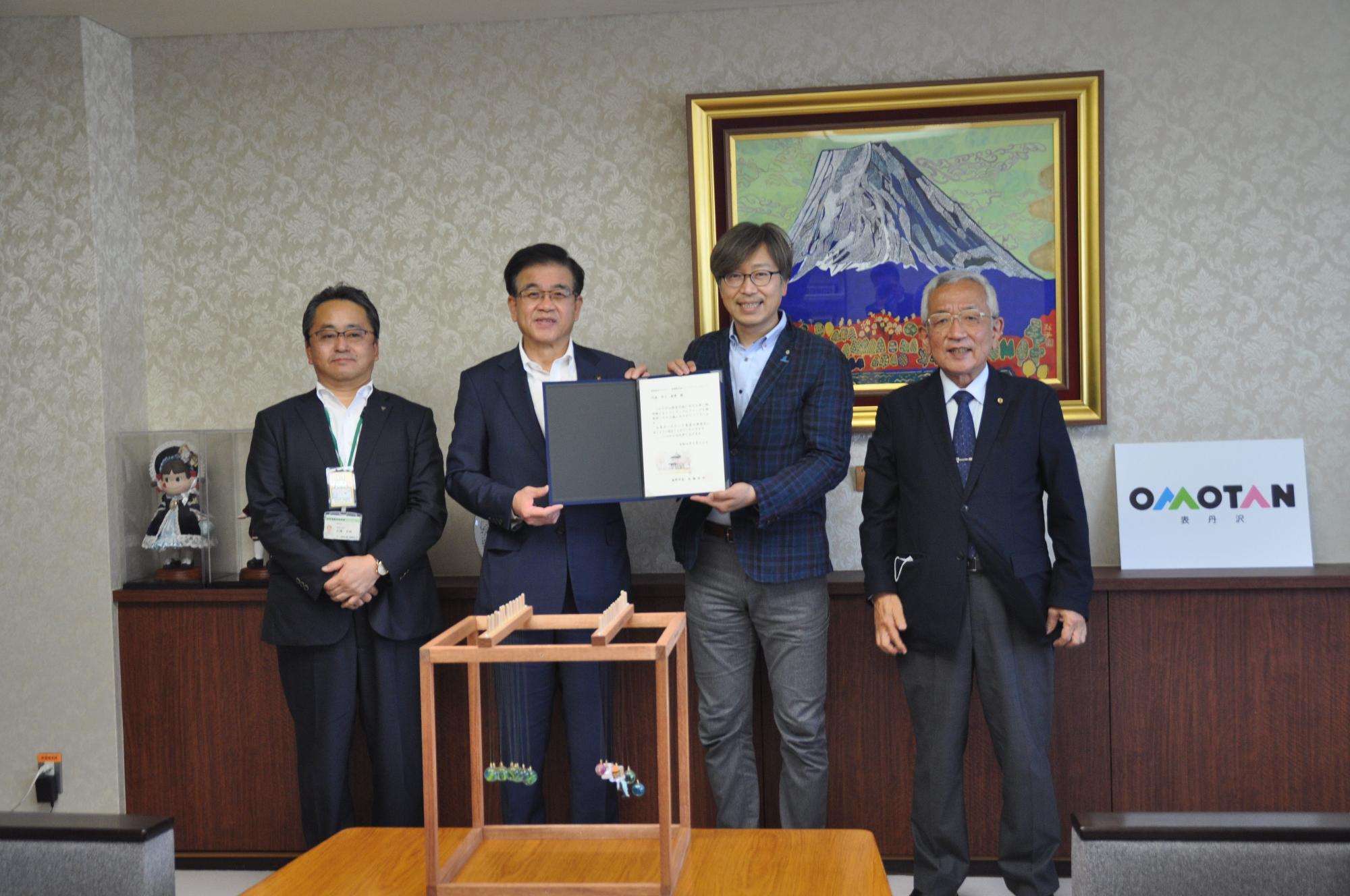 ペンデュラムウェーブが置かれたテーブルの前で高橋市長と井上代表が証書ホルダーを一緒に持ち、2人の両側に関係者の男性2名が並んで記念撮影をしている写真