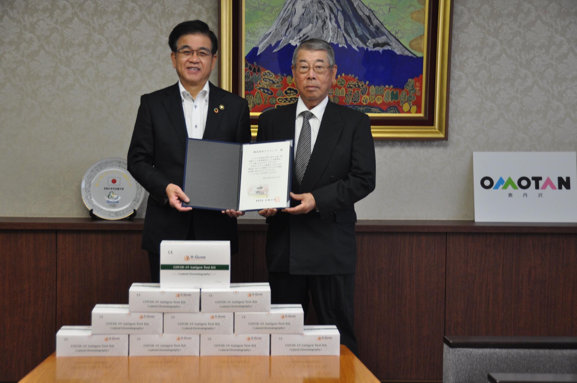 テーブルの上に積み重ねられた抗原検査キットを前に、高橋市長と佐藤代表取締役が一緒に証書ホルダーを持ち記念撮影をしている写真
