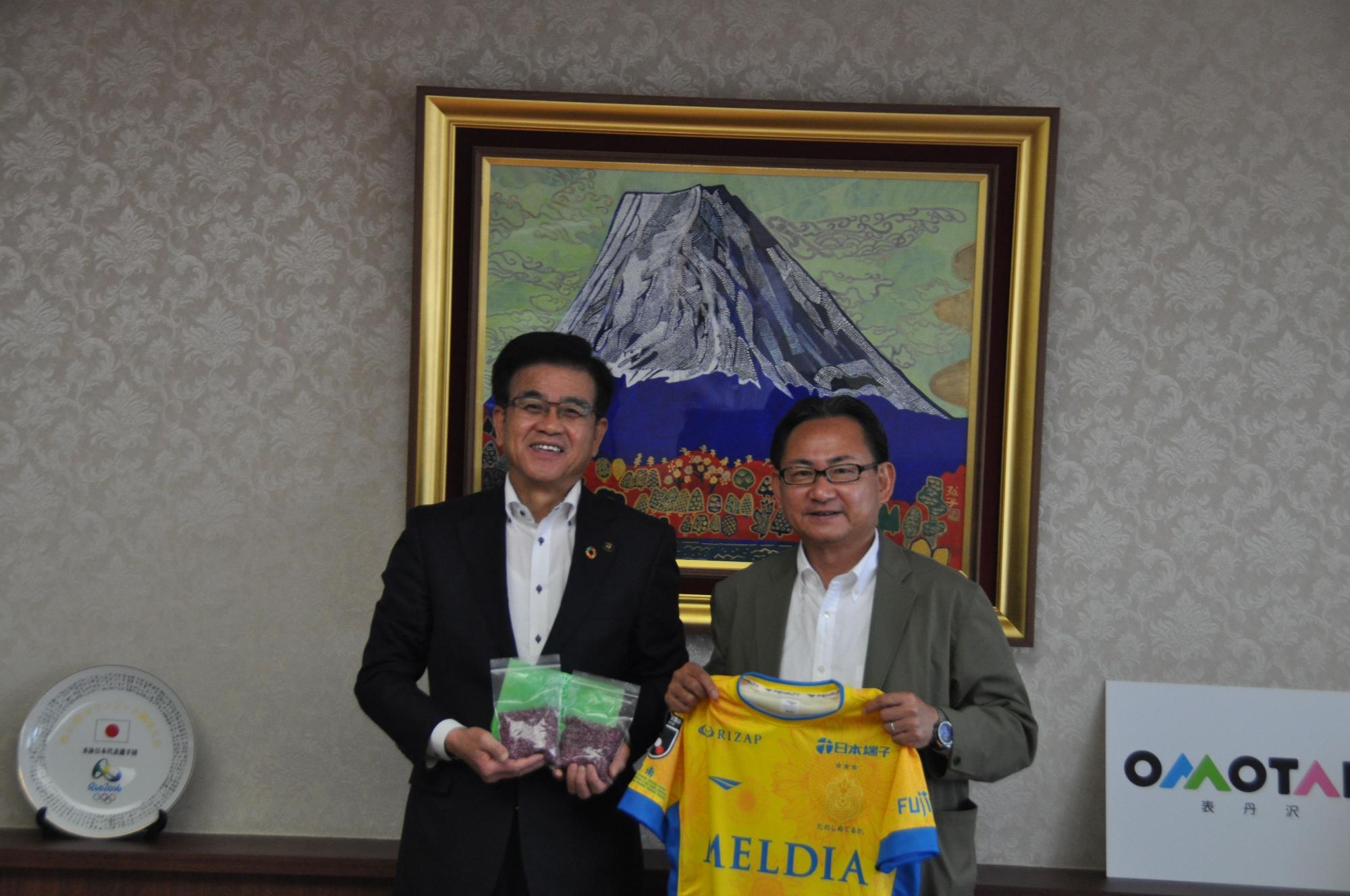 高橋市長とスーツ姿の男性が並んで立ち、高橋市長はヒマワリの種が入ったパックを、もう1名は「MELDIA」と書かれた黄色のユニフォームを持ってカメラに笑顔を向けている写真