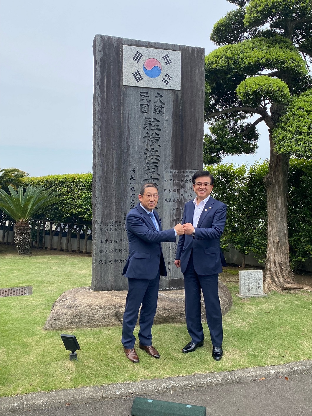 尹総領事と高橋市長が韓国国旗の付いた大きな石碑の前で拳を合わせ、笑顔でカメラにポーズをとっている様子を写した写真