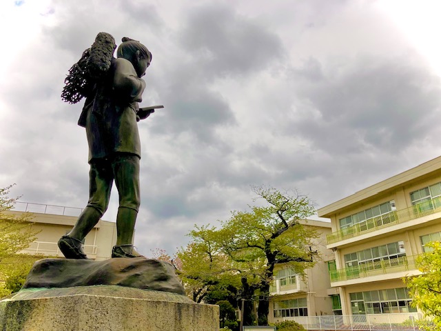 背中に柴を背負いながら本を読んでいる二宮金次郎の銅像が校庭に建ち、その背景には木々の緑と三階建ての校舎が写っている写真