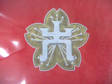 赤色の背景の中央に、金色の縁取りと薄い金色で描かれた桜の花びら5枚の上に、白い線で「大」の字をモチーフにしたデザインが描かれた校章の写真