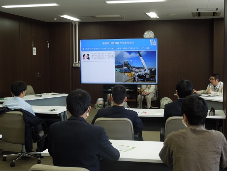 スーツ姿の参加者たちがマスクを着用してスクリーンのほうを向いており、男性が右奥でマイクを持って話している様子の写真