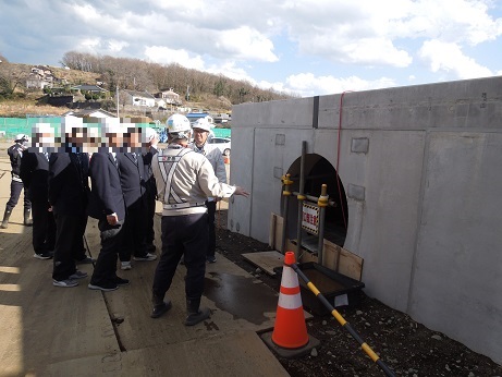 白いヘルメットと反射材付きの作業服を着た作業員が、数名のスーツ姿の男性たちに対し、新設されたコンクリート製の水路の構造物について説明している様子の写真
