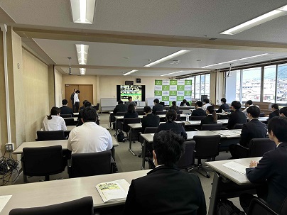 横長の会議室で、多数の参加者が前後左右に並んだ机と椅子に座り、プロジェクターに資料が映し出され、説明会が行われている様子の写真