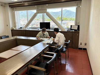 大きな窓際にある会議室で、2人の男性が座って資料を広げ、説明を聞いている様子の写真