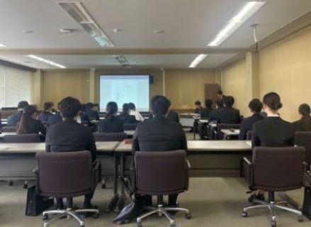 横長の会議室で、多数の参加者が前後左右に並んだ机と椅子に座り、プロジェクターに資料が映し出され、説明会が行われている様子の写真