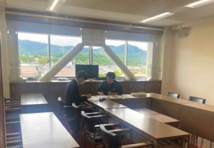 大きな窓際にある会議室で、2人の男性が座って資料を広げ、説明を聞いている様子の写真
