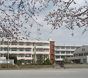 手前に広いグラウンドがあり、白色の4階建ての校舎が建ち、右側に体育館がある南中学校の外観写真