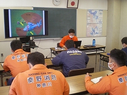 オレンジ色の救助服を着た数人の隊員が教室で真剣に座学を受けており、講師がPCを操作しながらプロジェクターでヘリコプターの図解を映している写真