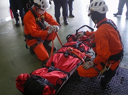 ヘルメットとハーネスを装着し、オレンジ色の救助服を着た二人の救助隊員が、ストレッチャーに負傷者を固定する実技訓練をしている様子の写真