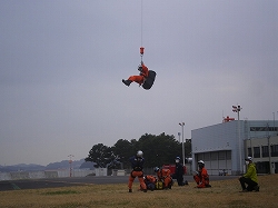屋外の訓練施設で、オレンジ色の救助服を着た隊員がヘリコプターから吊り下げられたワイヤーで空中に吊り下げられており、他の隊員がその様子を地上で見守っている訓練風景の写真
