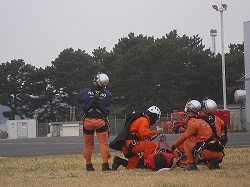 オレンジ色の救助服と白いヘルメットを着用した4人の隊員が、担架を囲んで膝立ちになり、連携している訓練風景の写真