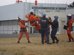 屋外の広場で、オレンジ色の救助服を着た救助隊員がヘリコプターから吊り下げられた担架を地上で待機する救助隊員と連携して訓練を行っている写真