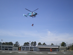 青と白の機体のヘリコプターが訓練施設の上空で、機体から長く伸びたワイヤーでオレンジ色の救助服を着た隊員が吊り下げられている救助訓練の様子の写真