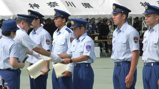 紺色の制帽をかぶり淡い水色のシャツと紺色のズボンを着用した若手の消防士たちが横一列に整列する中、向かい合いになり順番に表彰状を渡す男性の写真