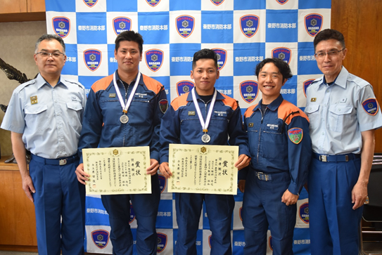 消防本部のロゴと「長野市消防本部」の文字が描かれた青い壁の前で、表彰状と首から下げたメダルを手に、オレンジと紺色の活動服姿の三名の消防士が左右に立つ制服姿の二名の幹部と共に記念撮影をしている写真