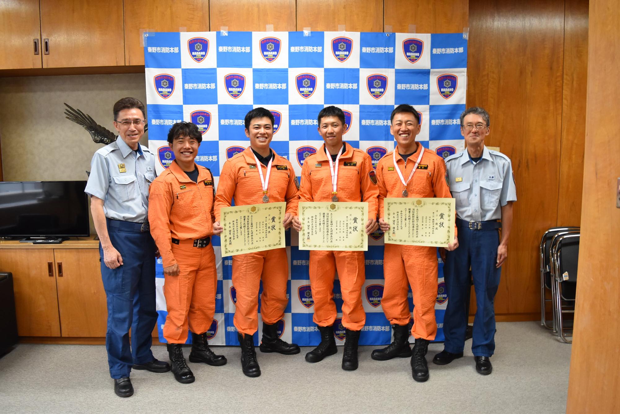 バックパネルの前で、メダルを首にかけたオレンジ色の活動服の4人の隊員が、表彰状を持って両脇の制服姿の2人の男性とともに並んで笑顔で立っている、集合写真