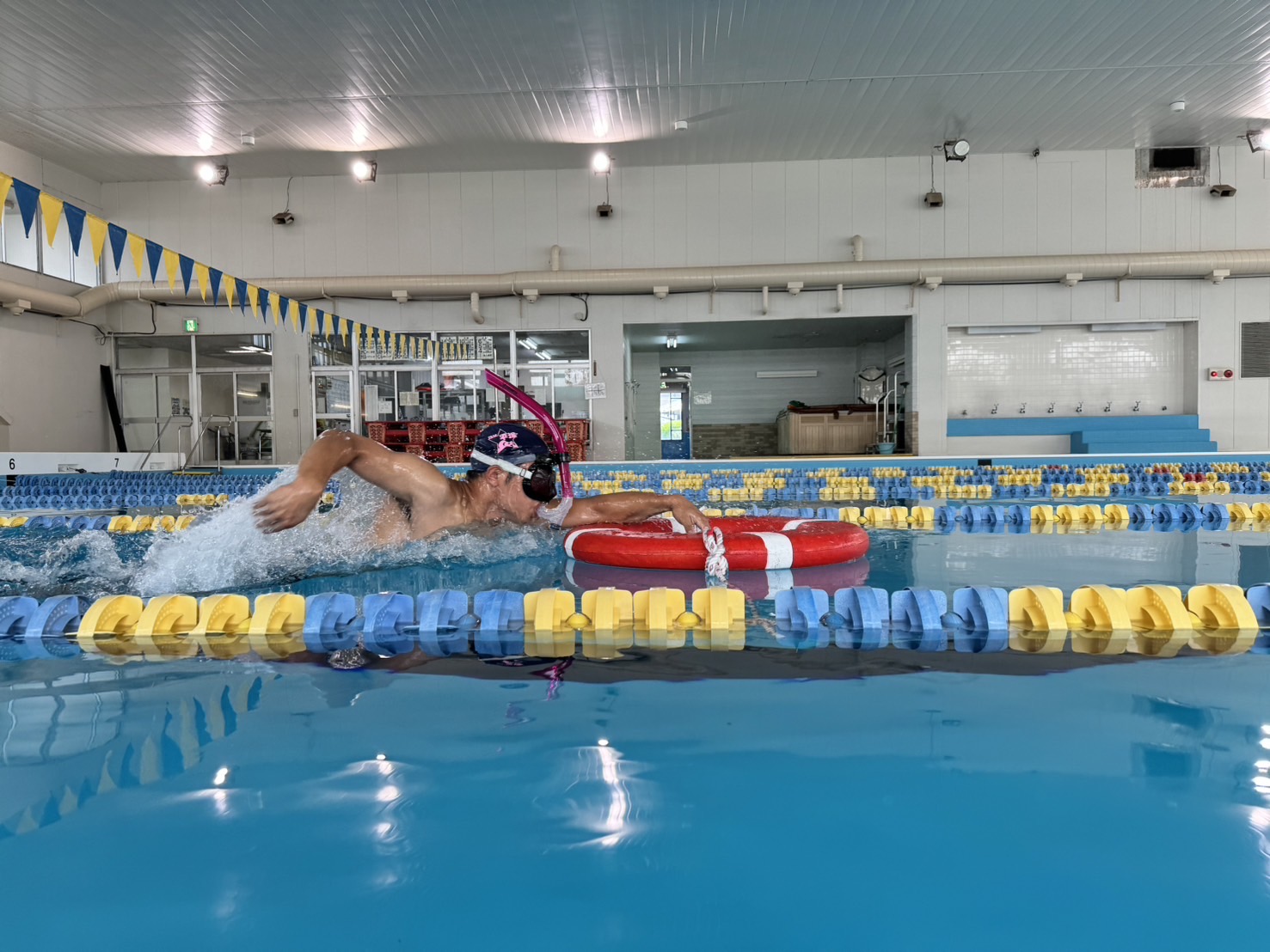 シュノーケルと水泳帽を着用した男性が、赤い浮き輪を右手に持ってクロールで泳いでいる様子を、屋内のプールサイドから撮影した写真