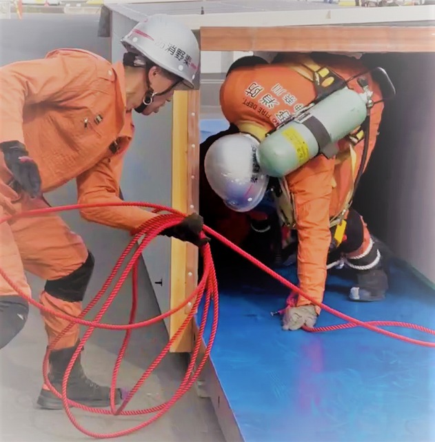 オレンジ色の防火服にヘルメットを着用した2人の救助隊員が、空気呼吸器を着装し訓練施設らしき狭い空間で活動しており、手前の隊員が赤いロープを捌いている写真