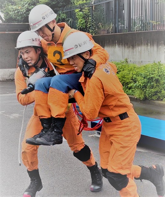 オレンジ色の防火服とヘルメットを着用した2人の救助隊員が、負傷者を模したもう一人の隊員を2人がかりで抱えて救助している様子を屋外で撮影した、救助訓練の写真