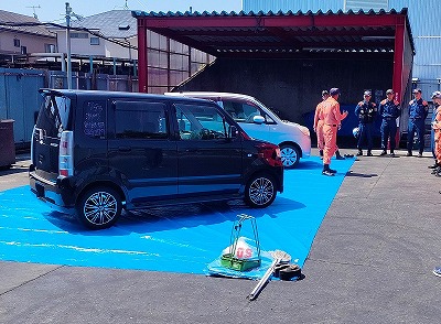 オレンジ色の活動服を着た隊員が数人の紺色の活動服の隊員に向かって話をしており、手前に黒と白の軽自動車が青いビニールシートの上に停車している写真