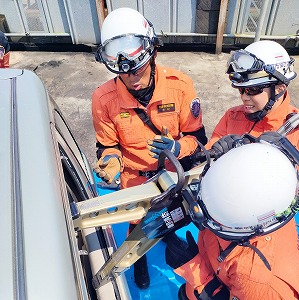 明るいオレンジ色の活動服と白いヘルメットを着用した3人の救助隊員が、大型油圧救助器具を使い、自動車のドアのこじ開け訓練を実施している様子の写真