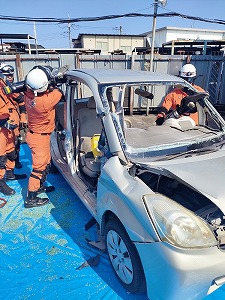 白いヘルメットとオレンジ色の活動服を着用した複数の救助隊員が、フロントガラスやルーフの一部が切断された銀色の軽自動車を用いて訓練を行っている様子を捉えた写真