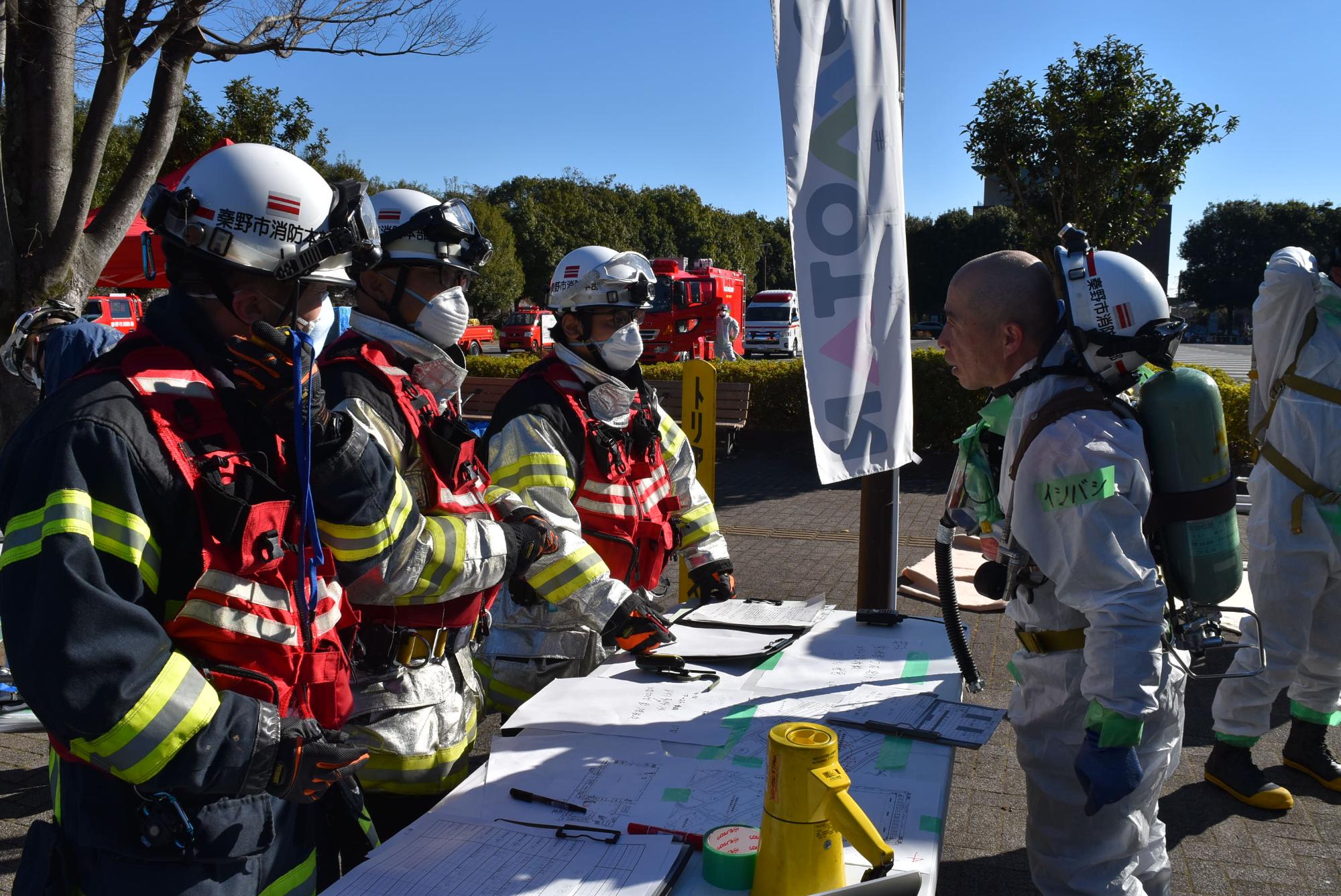 白いヘルメットやマスク、防火服、化学防護服などを着用した隊員たちが、屋外の机を挟んで打ち合わせをしている様子を捉えた、防災訓練の写真