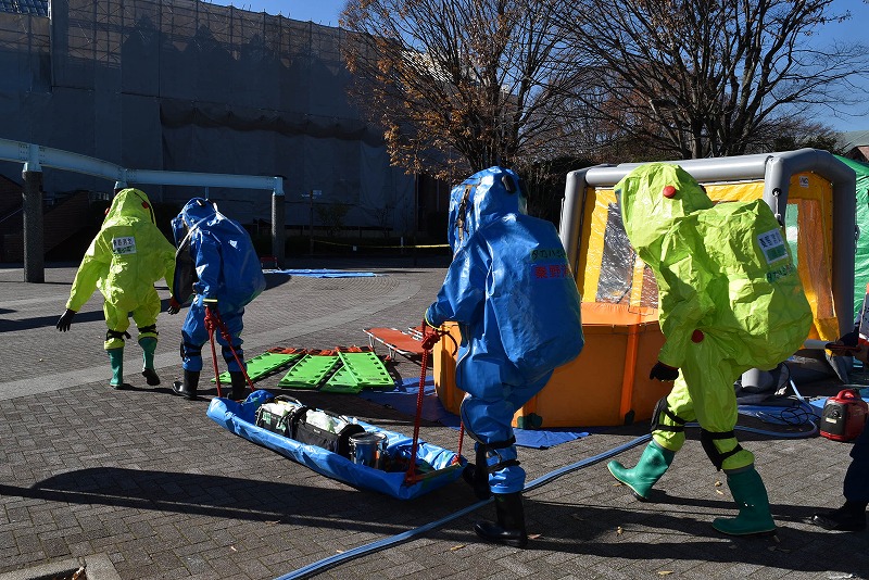 青と蛍光黄色の化学防護服を着用した隊員たちが、除染テントのような構造物近くで担架の上に積載された機材を引っ張っている写真
