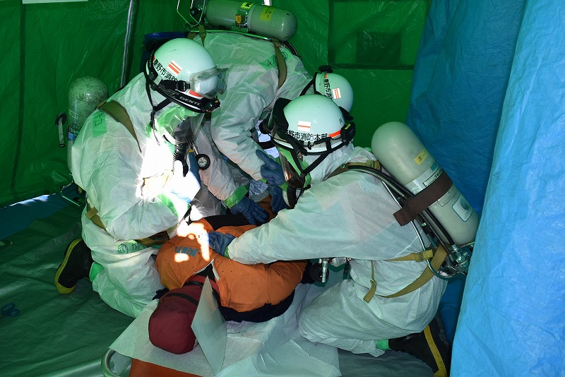 密閉された青いテントの中で、白い化学防護服と呼吸器を装着した3人の隊員が、担架の上のオレンジ色の服を着た人形に対応している救助訓練の写真