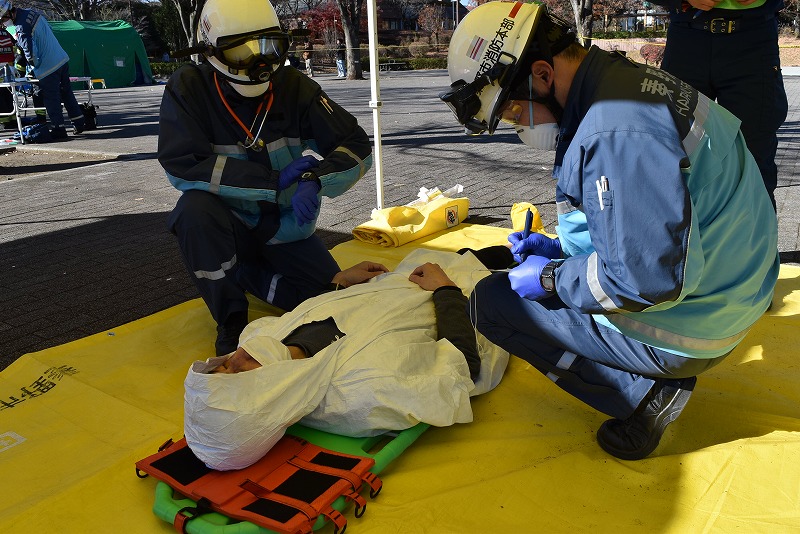 屋外の黄色いシートの上で、白い防護シートに包まれた人をストレッチャーに乗せた状態で、2名の隊員が対応している救助訓練の様子の写真