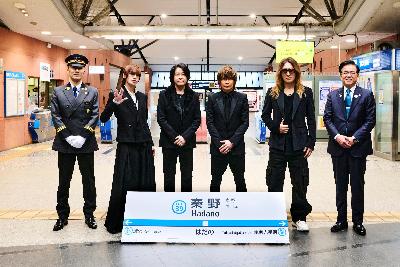 11月27日（木曜日）に開催した「秦野駅列車接近メロディー導入記念セレモニー」に出席した、左から、栗林範之秦野駅長、ルナシーのSUGIZOさん、RYUICHIさん、INORANさん、Jさん、高橋昌和市長が、改札前に横一列で並び、その前に秦野駅の駅名標が立てかけられている集合写真
