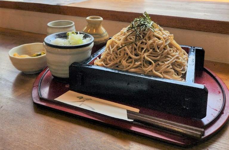 木目調のテーブルの上で、赤いお盆に乗せられたざる蕎麦と、つゆの入った徳利、薬味の入ったそば猪口が揃っている様子の写真