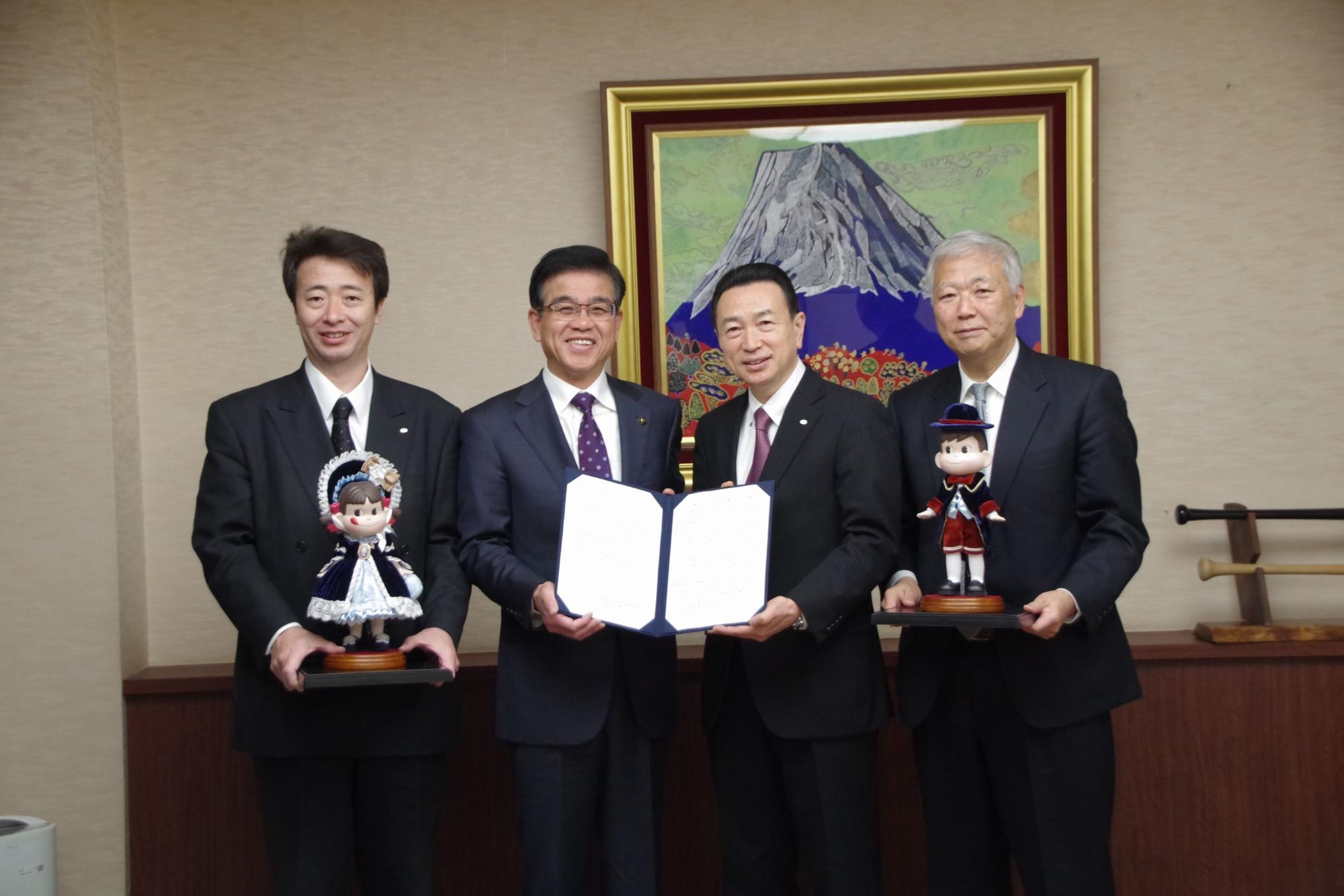 市長と中央の男性が契約書を持ち、両端に立った男性2人が西洋風の衣装を着たペコちゃん、ポコちゃんの人形それぞれ持って並んで立っている記念写真