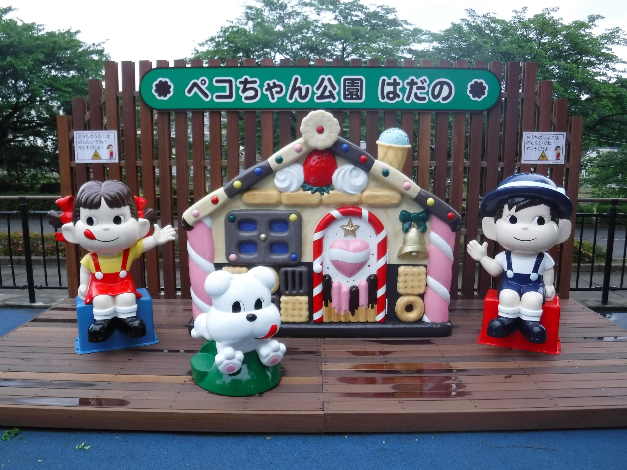 「ペコちゃん公園 はだの」と書かれた看板の下、中央のお菓子の家を挟んで、赤いオーバーオール姿のペコちゃんと青いオーバーオール姿のポコちゃん、手前に白い犬のキャラクターが配置された可愛らしいオブジェの写真