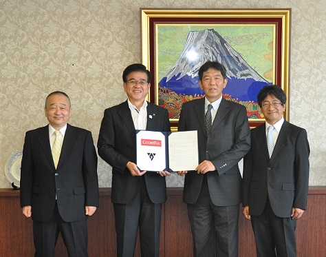 スーツを着た市長と3人の男性が並んで立ち、市長と中央の男性が契約書持って写した記念写真