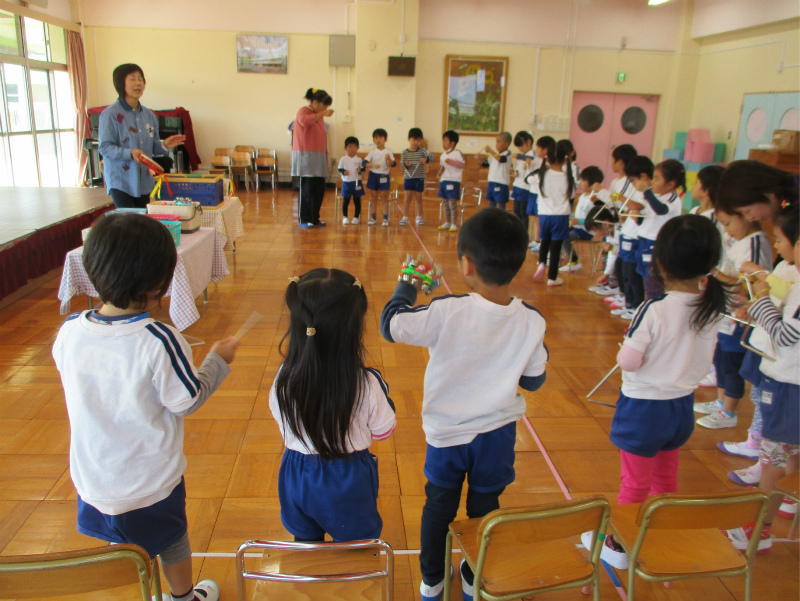 体操服を着た多くの園児たちが楽器のような道具を手に持って並び、2人の先生に教わりながら音楽を演奏している様子の写真