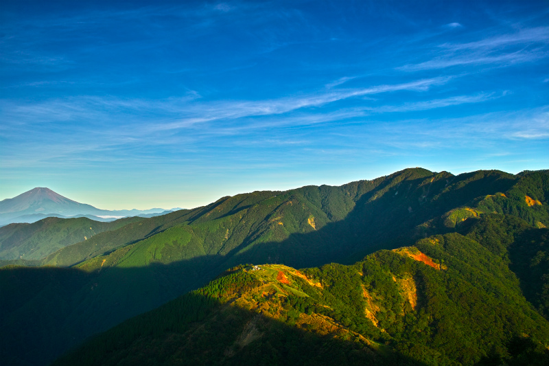 山脈の稜線に沿って光と影がくっきりと分かれ、遠景に富士山が霞んで見える美しい風景写真