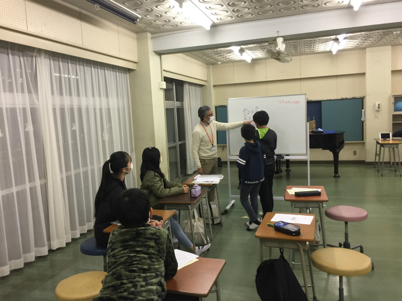 男性教師と2人の男子生徒がホワイトボードの前に立ち、他の生徒たちが机に向かって真剣に授業を受けている様子の写真