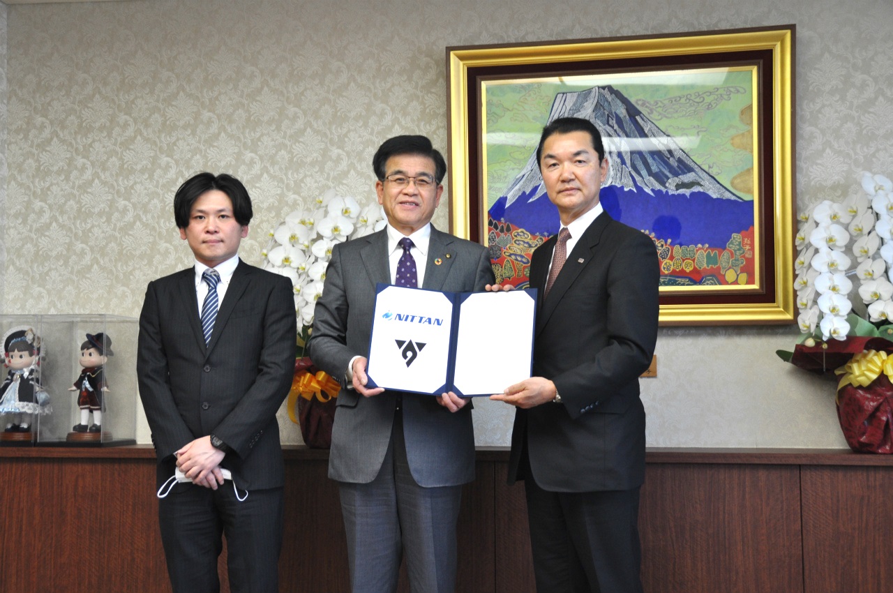 スーツ姿の市長の両側に1人ずつ男性が並び、市長と右側の男性が「NITTAN」のロゴが入った契約書を持って写した記念写真