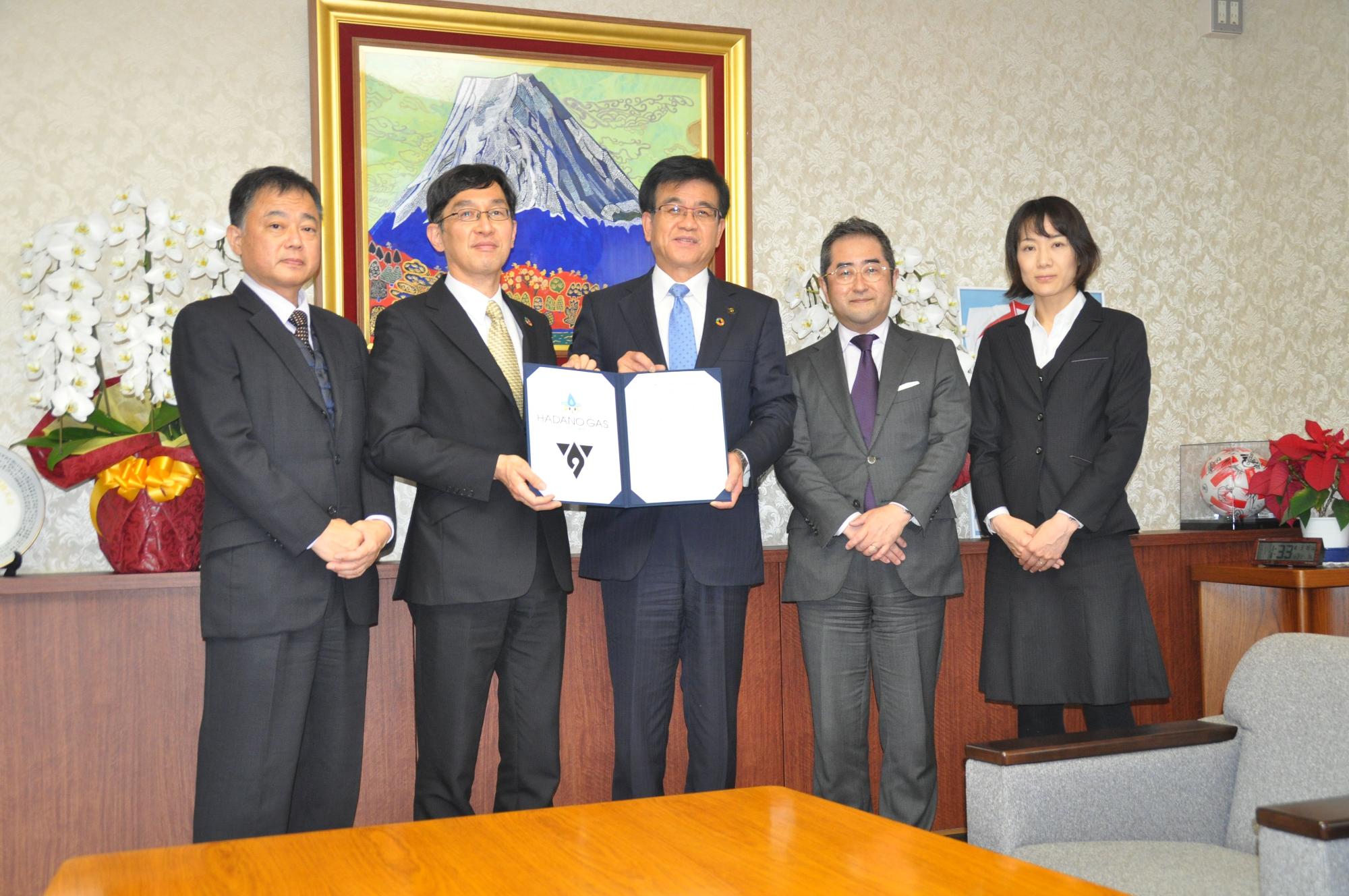 スーツを着た市長と関係者が、中央の男性が持つ「はだのガス」のロゴが入った契約書を囲んで立った並んだ記念写真
