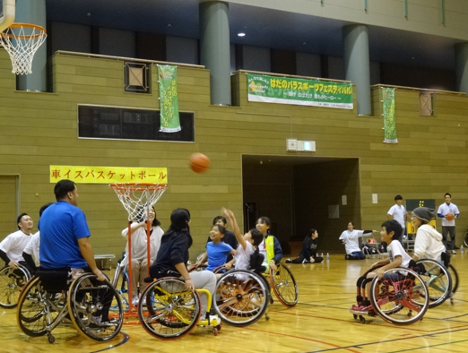 壁に「車いすバスケットボール」と書かれた黄色の横断幕が貼られた体育館のバスケットコートで、車いすに乗った人々がバスケットボールを楽しんでいる様子の写真
