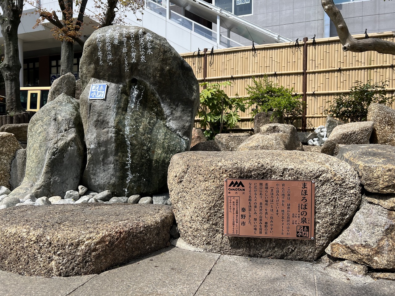 竹柵が設置された敷地内に、中央から水が流れている岩の隣に設置された「まほろばの泉」と書かれた石碑の写真