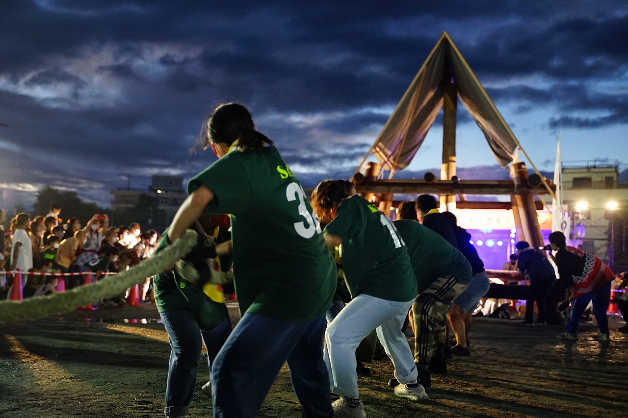 日が暮れかけた夜祭の会場で、三角屋根の櫓の中央に設置された大きな柱に結んだ大きな綱を、お揃いの緑色のTシャツを着たチームが全力で引いているジャンボ火起こし綱引きコンテストの様子を撮影した写真