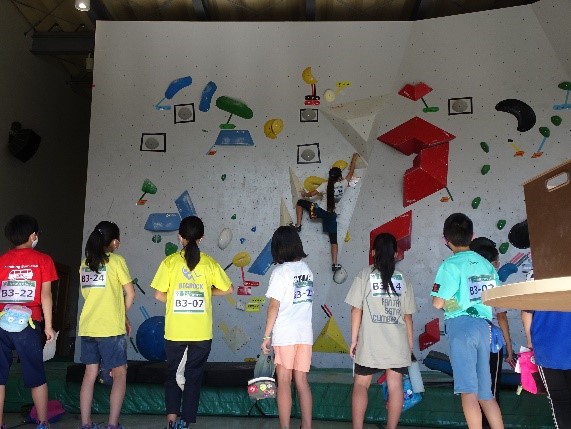 壁のいたる所にカラフルな人工の石や突起物が取り付けられているボルダリングの競技会場で、ゼッケンをつけた複数の子供たちが登っている友達を真剣に見守っている写真